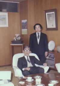椅子に座る加賀市長と横に並んで立つスーツを着た男性。